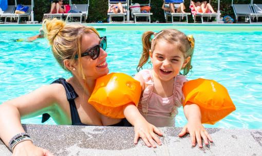 Madre e figlia sorridenti in piscina con braccioli arancioni.