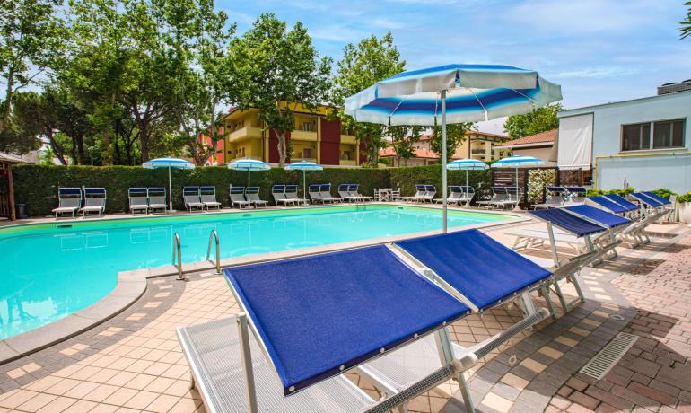 Piscina all'aperto con lettini e ombrelloni blu, circondata da alberi.