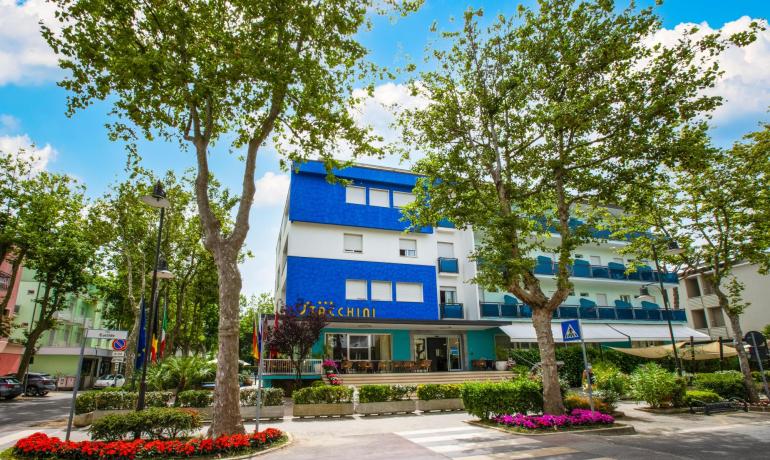 Hotel moderno con facciata blu, circondato da alberi e fiori colorati.