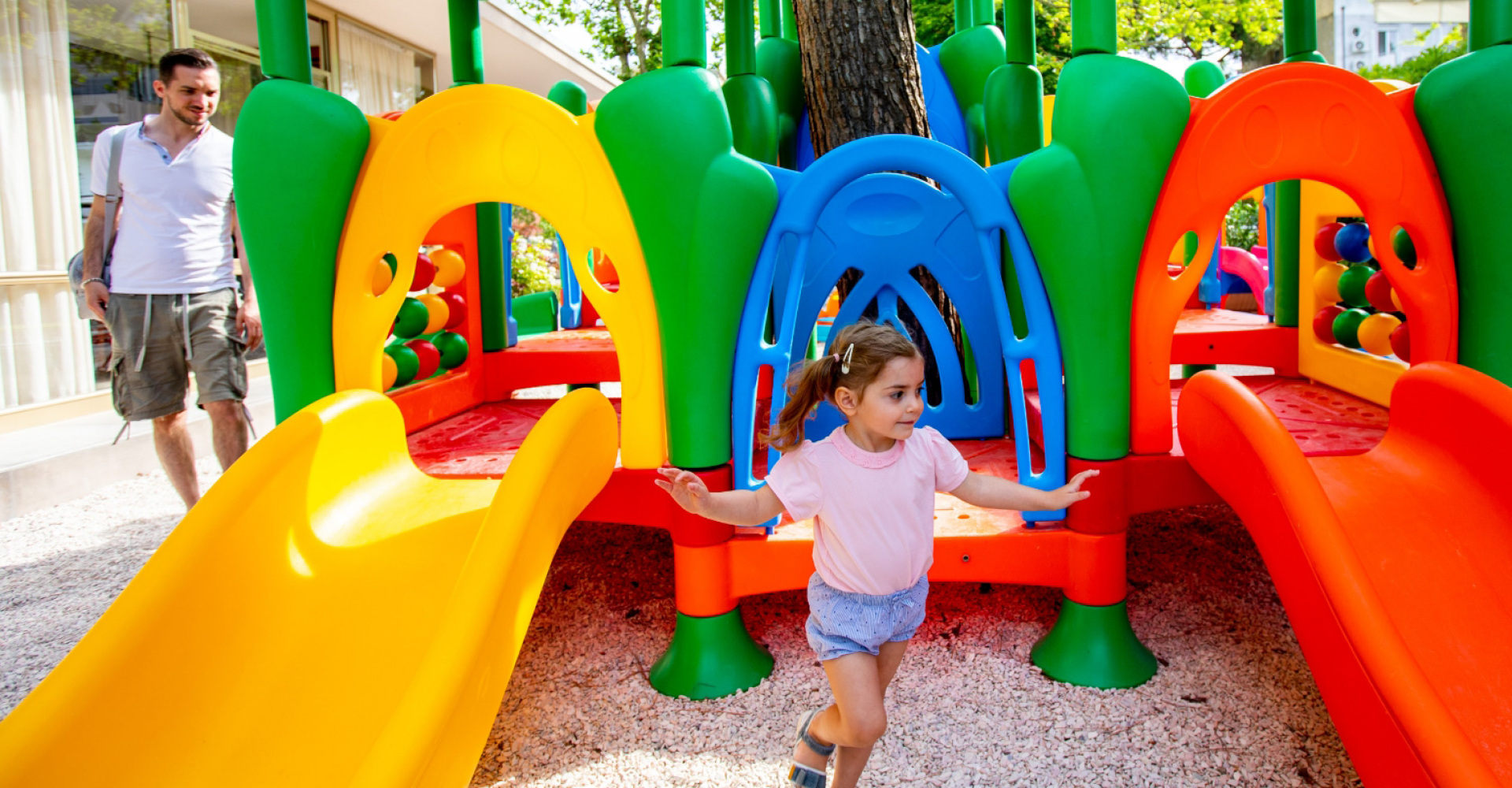 Bambina gioca su uno scivolo colorato, uomo osserva in un parco giochi.