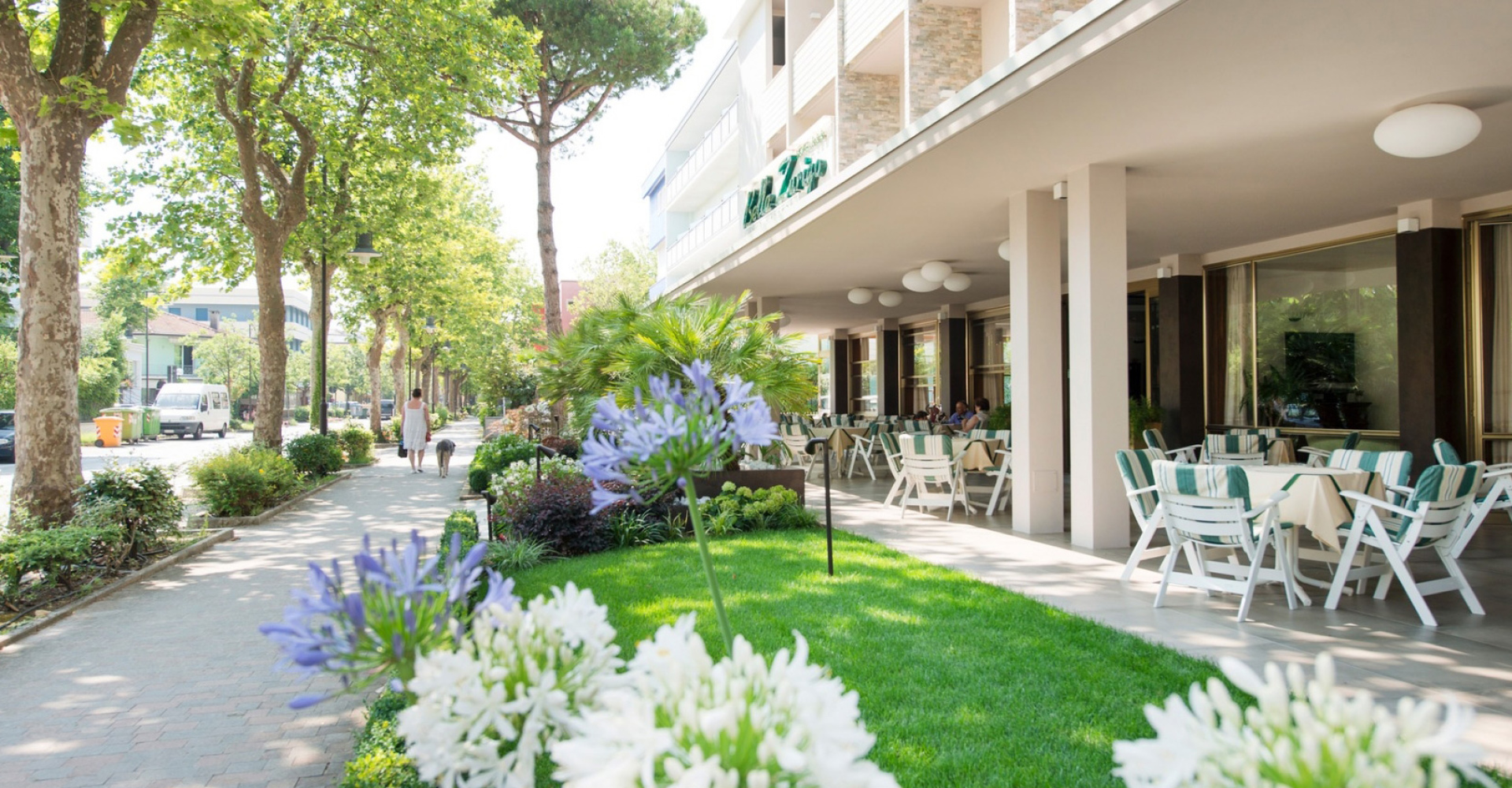 Giardino esterno con tavoli, sedie bianche e fiori, lungo un viale alberato.