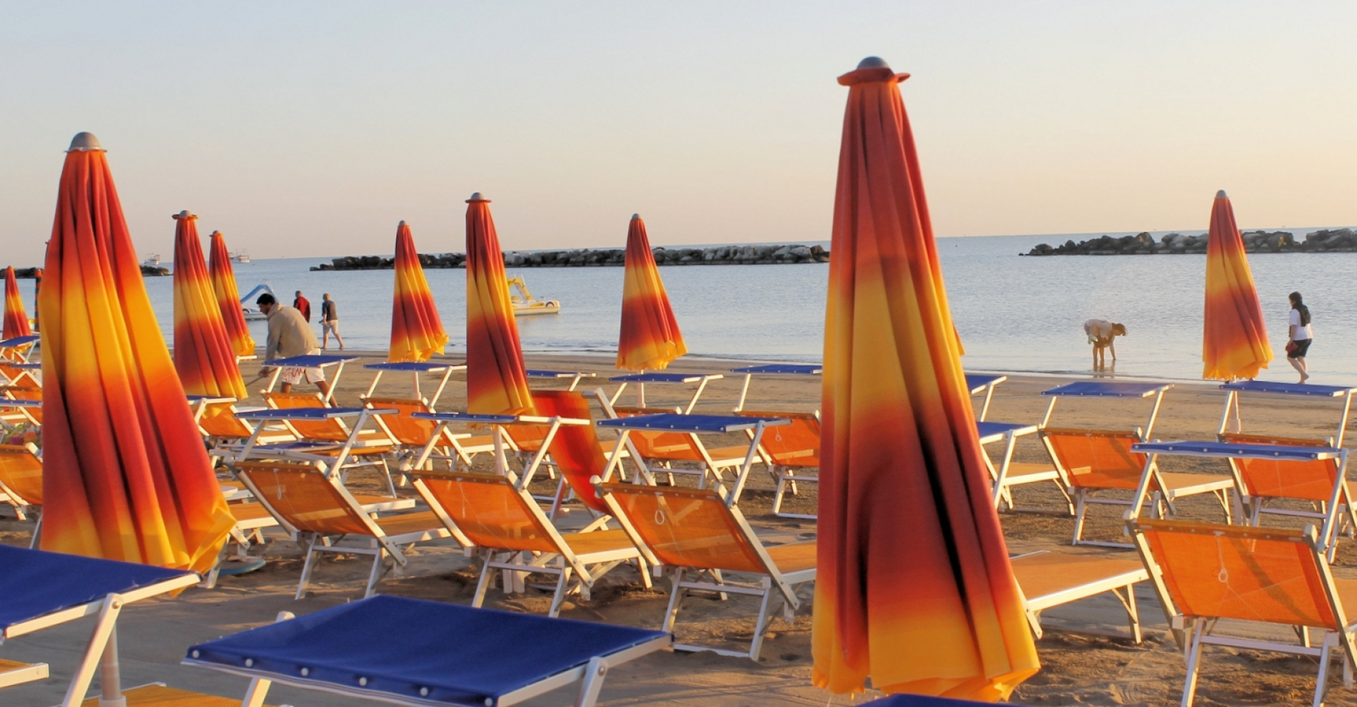 Spiaggia al tramonto con ombrelloni colorati e lettini vuoti, persone passeggiano sulla riva.