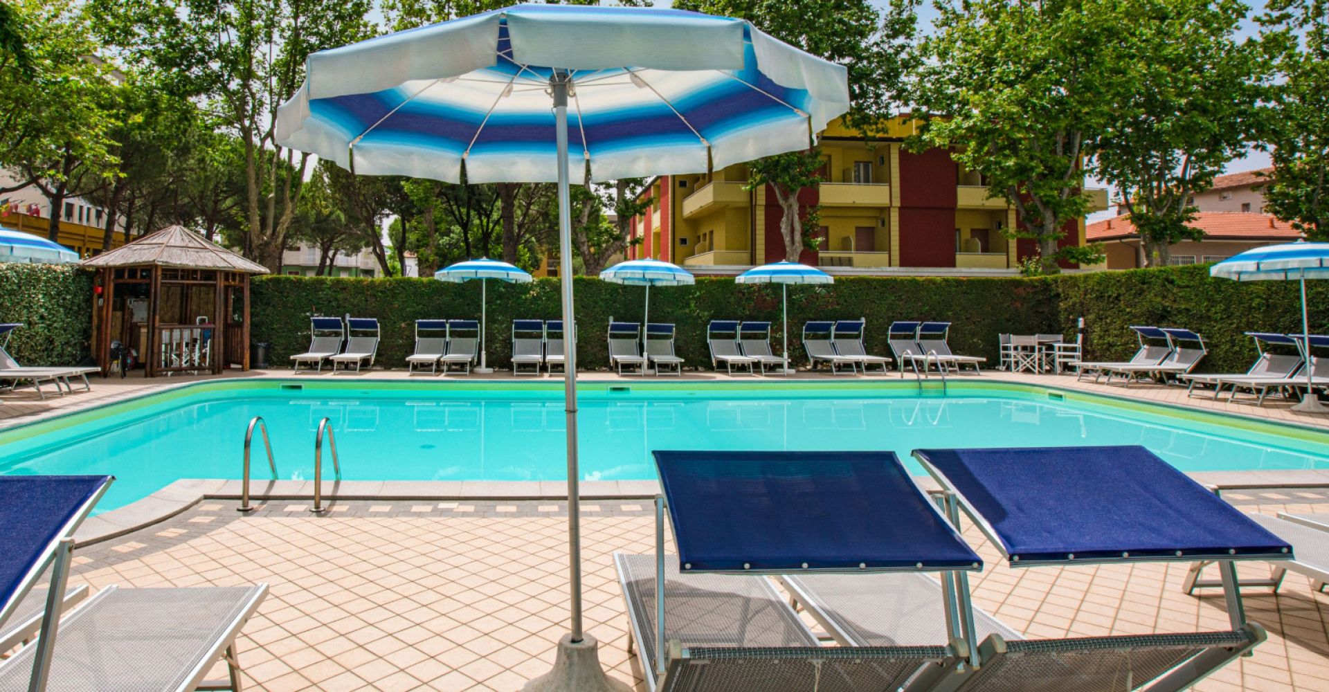 Piscina all'aperto con lettini e ombrelloni, circondata da alberi e siepi.