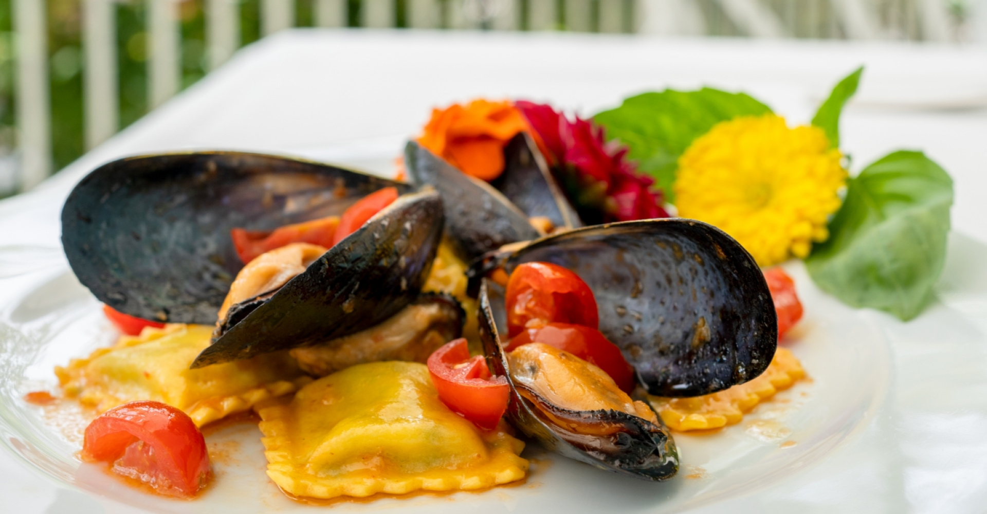 Ravioli con cozze, pomodorini e decorazione floreale su piatto bianco.