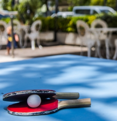 Racchette da ping pong e pallina su un tavolo all'aperto, sfondo sfocato.