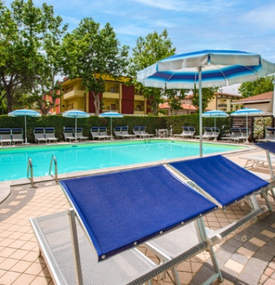 Piscina all'aperto con lettini blu e ombrelloni, circondata da alberi e edifici.