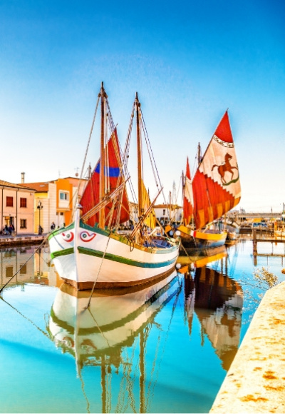 Barche colorate con vele rosse in un canale riflettente, vicino a edifici storici.