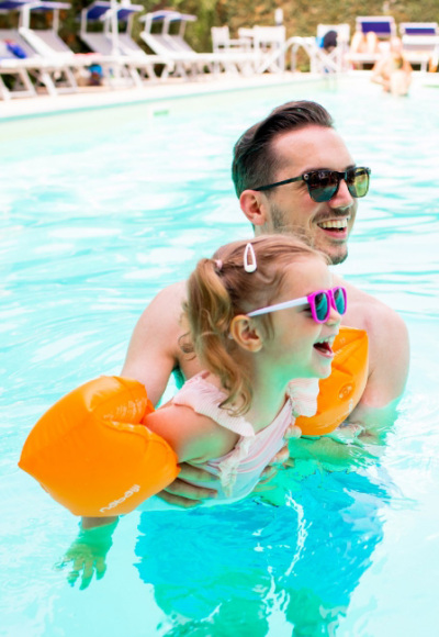 Papà e figlia sorridenti nuotano insieme in piscina con occhiali da sole.