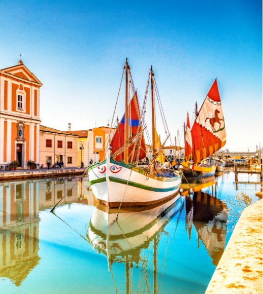 Barche colorate con vele rosse in un canale riflettente, vicino a edifici storici.