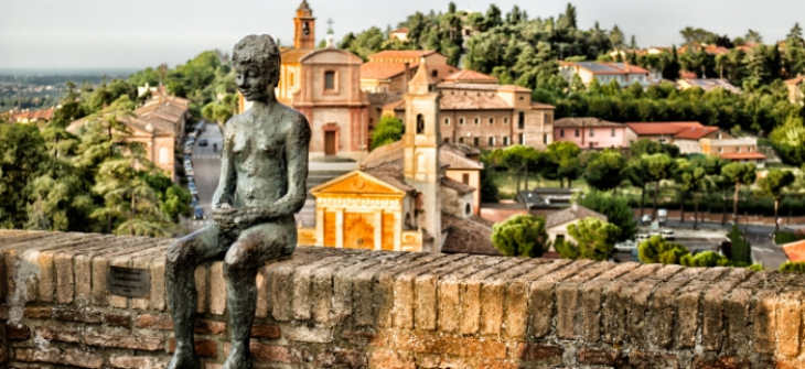 Statua di bronzo su un muretto, con vista su un borgo italiano.