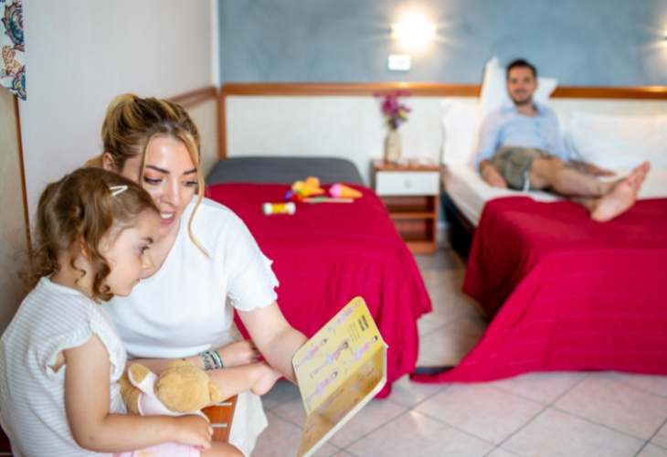 Famiglia in hotel: mamma legge al bambino, papà si rilassa sul letto.