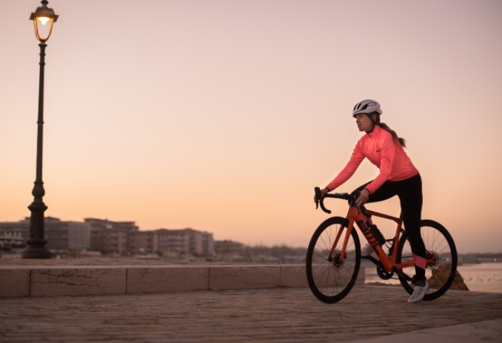 Ciclista al tramonto, indossa abbigliamento sportivo e casco, su una strada costiera.