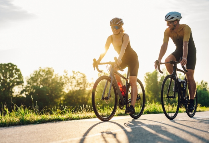 Due ciclisti pedalano su strada al tramonto, indossando abbigliamento sportivo e caschi.