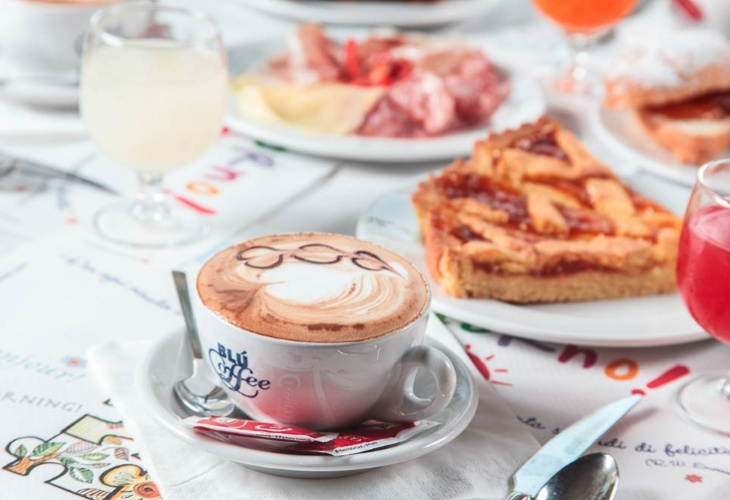 Colazione italiana con cappuccino, crostata e affettati su una tavola imbandita.