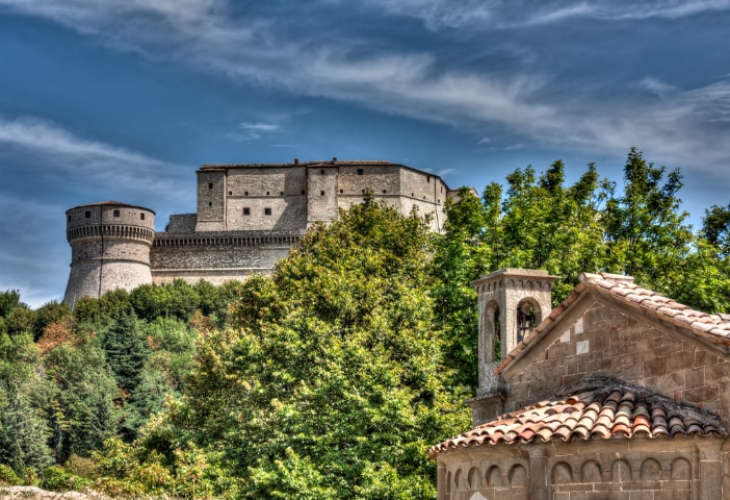 Castello medievale tra alberi e cielo azzurro, con una piccola cappella in primo piano.