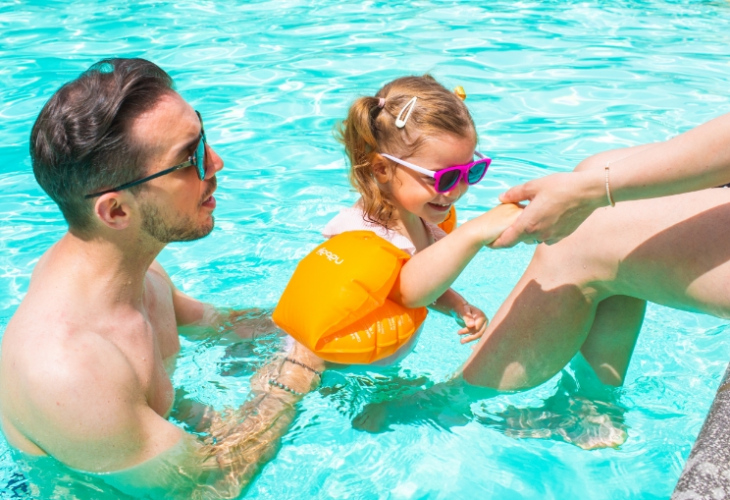 Bambina con occhiali da sole e braccioli arancioni gioca in piscina con la famiglia.