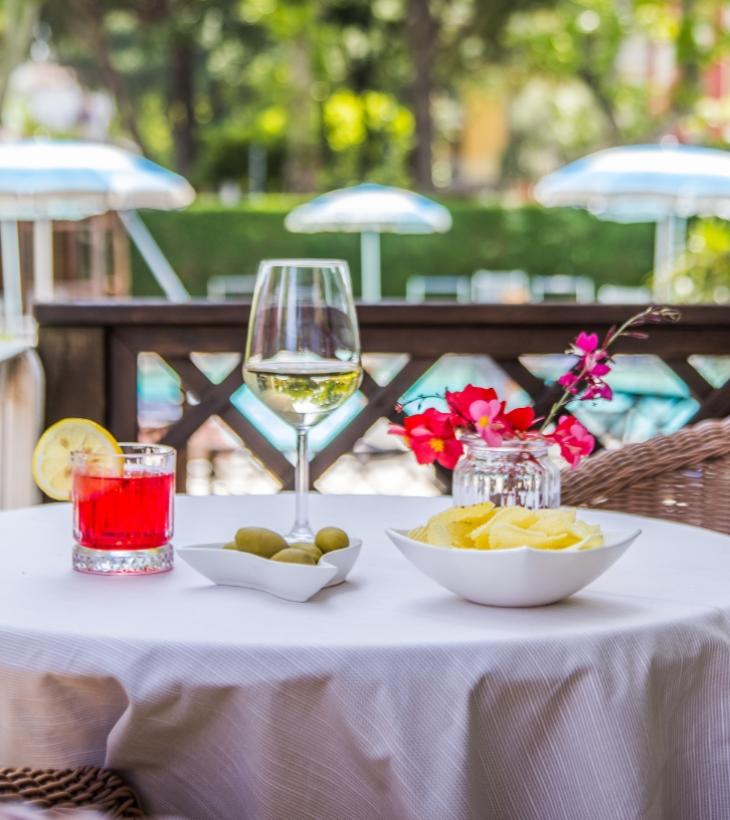 Tavolo con drink, olive, patatine e fiori, vista su ombrelloni in giardino.