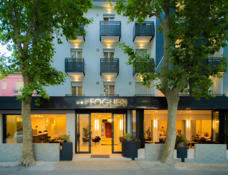 Hotel moderno con balconi e illuminazione calda, alberi davanti.