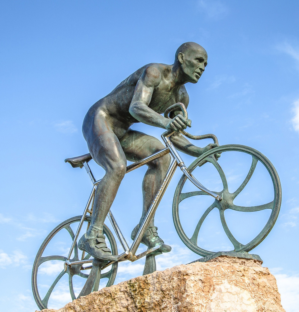 Statua di ciclista su roccia, in metallo, contro un cielo azzurro.