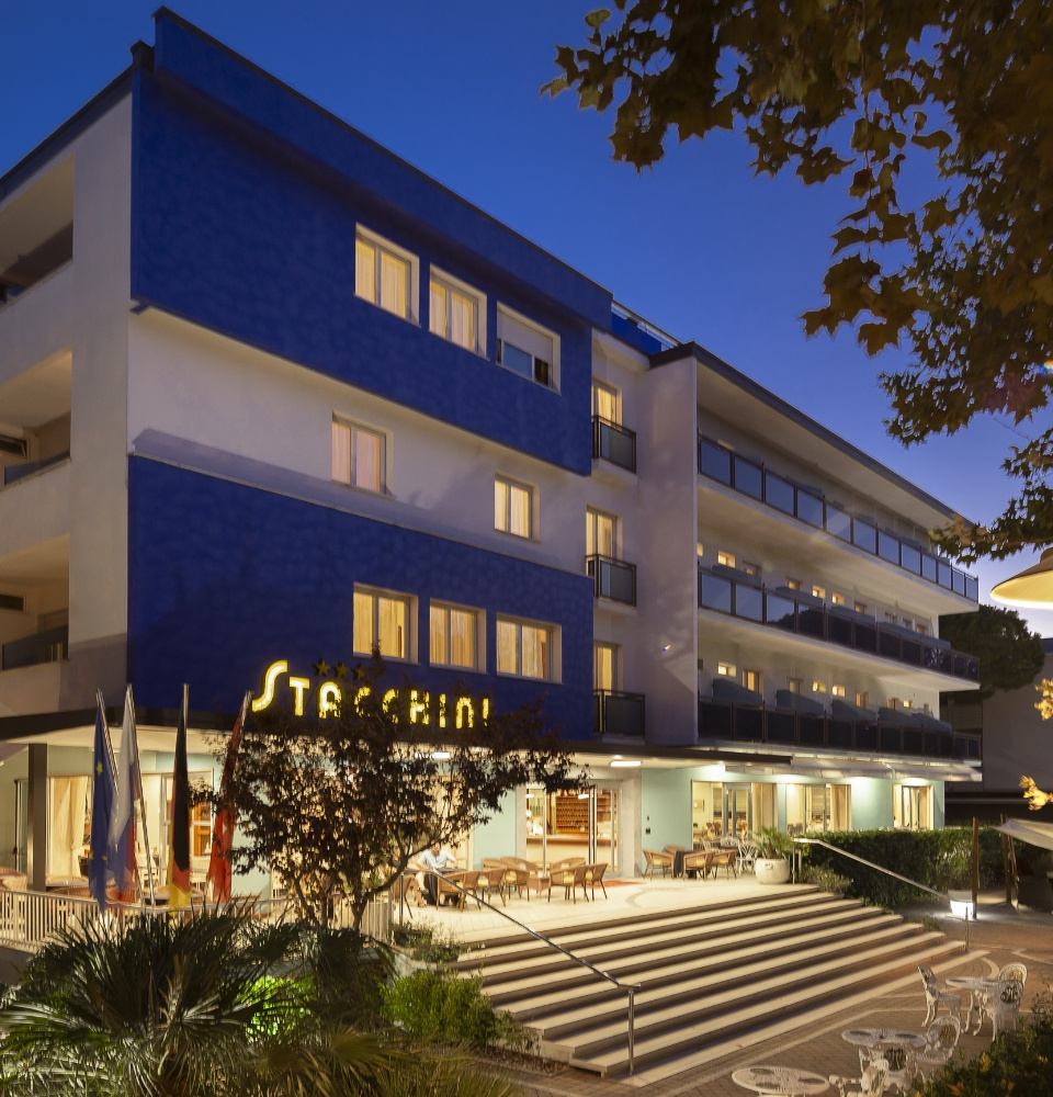 Hotel moderno con terrazze, scale d'ingresso e tavolini esterni, illuminato di sera.