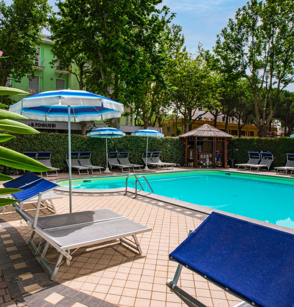 Piscina con lettini e ombrelloni, circondata da alberi e siepi verdi.