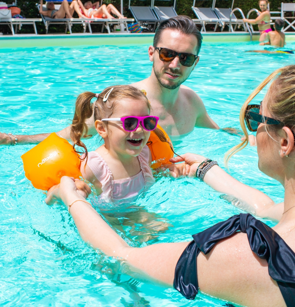 Famiglia in piscina, bambina con occhiali da sole e braccioli arancioni.