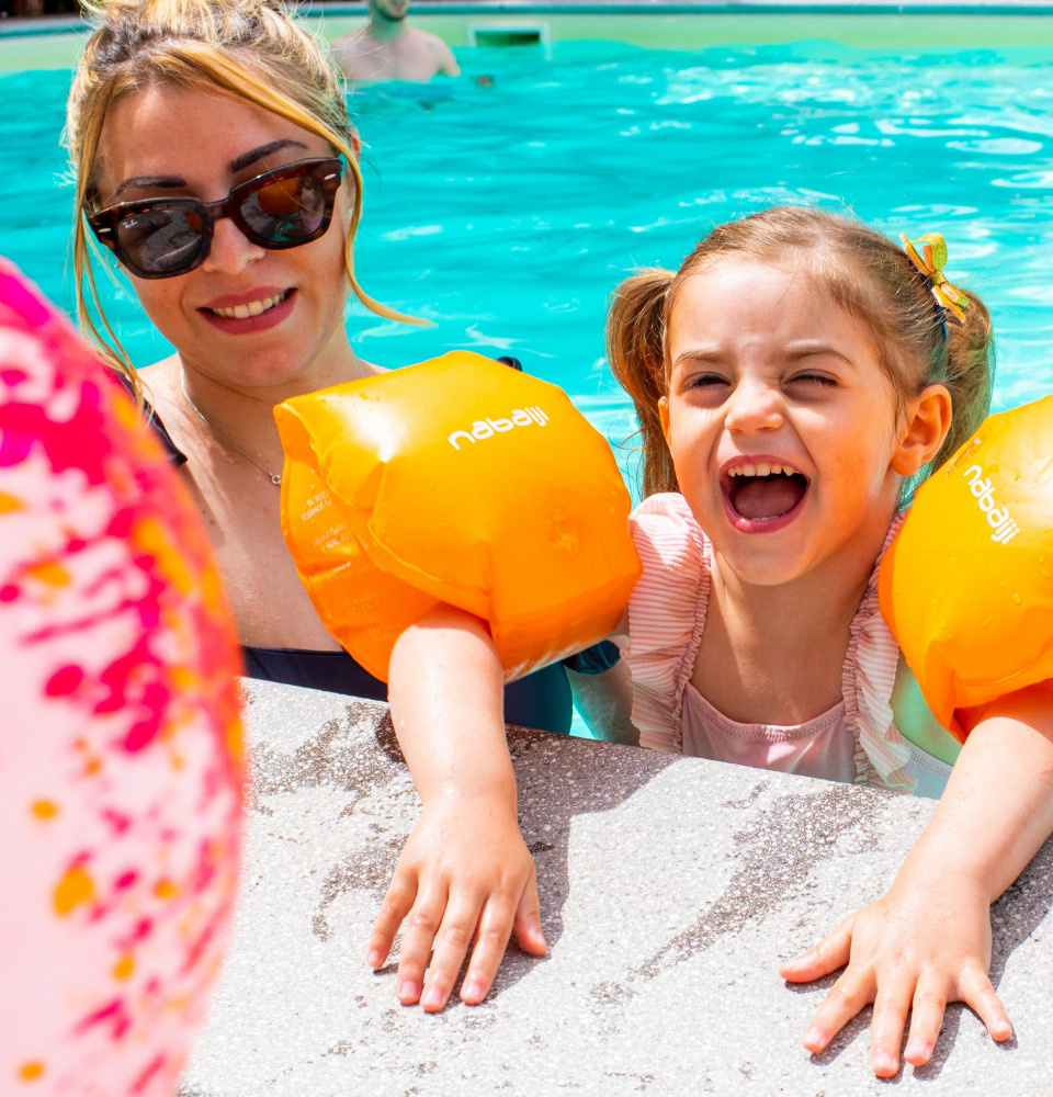 Bambina e donna sorridenti in piscina con braccioli arancioni, palla colorata in primo piano.